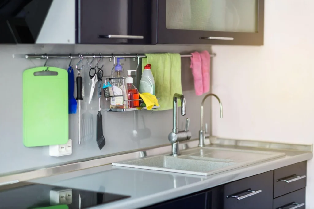 Organized kitchen sink area with stainless steel sink, faucet, and wall-mounted organizer with cutting board, utensils, towels, and cleaning supplies, perfect for clutter-free waste solutions with under cabinet garbage bins.