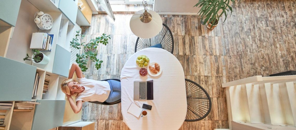 Top view of a woman relaxing in a bright compact dining area with a laptop and breakfast on the table, illustrating a tidy, clutter-free small home.